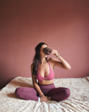 Woman in pink sports bra and maroon leggings sitting on a bed, drinking from a brown mug.